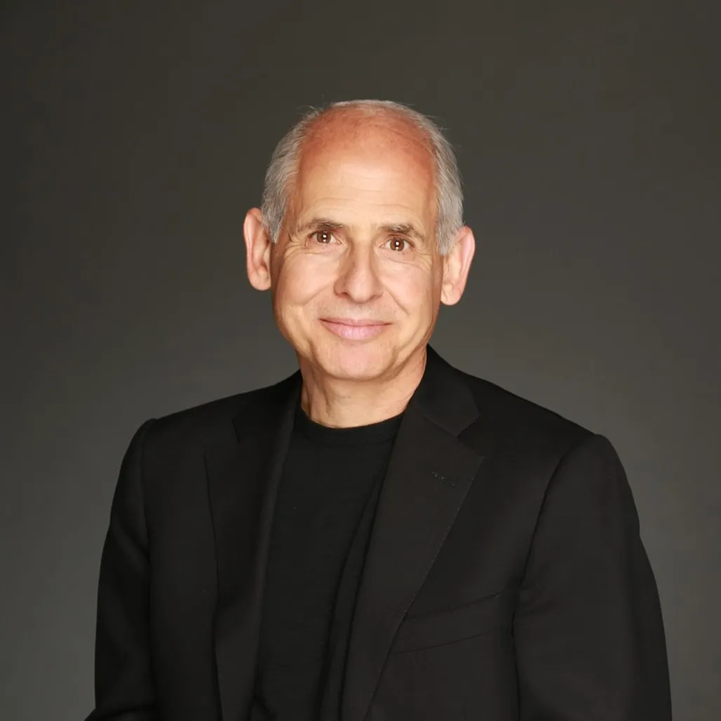 Smiling older man with short gray hair wearing a black blazer and black shirt against a dark background.